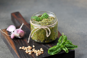 Pesto green sauce in a glass jar. On a wooden board next to a jar of garlic, basil greens and pine...