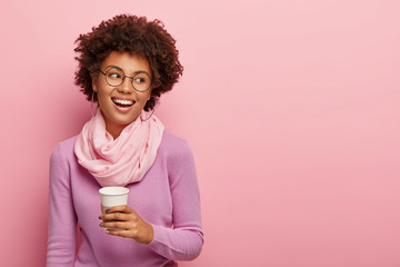 Positive relaxed curly female worker enjoys coffee break, has talk with colleague, holds disposable...