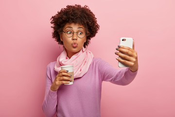 Young lovely woman with Afro haircut, keeps lips folded, blows kiss at camera of smartphone, takes...