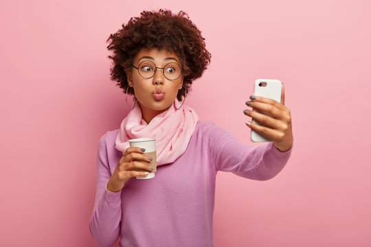 Young lovely woman with Afro haircut, keeps lips folded, blows kiss at camera of smartphone, takes selfie portrait, drinks takeaway coffee, wears stylish clothes, transparent glasses, isolated on pink