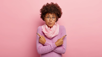 Wondered hesitant Afro woman points index fingers to different sides, cannot choose what to pick, has doubtful look, dressed in soft purple jumper, silk scarf on neck, decides what is better
