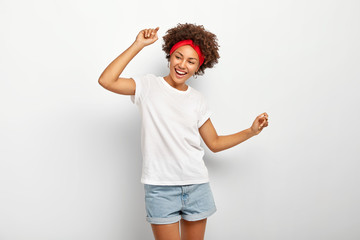 Carefree pleased teenage girl has fun, dances joyfully with raised arms, being entertained and amused, wears summer clothes, laughs happily, enjoys cool music, isolated on white wall, being on party