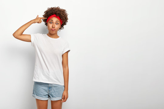 Crazy funny Afro girl shoots in temple with finger, gestures indoor, makes suicide gesture, has surprised face expression, dressed in casual summer wear, stands against white wall with copy space