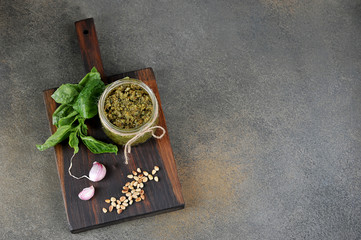 Pesto green sauce in a glass jar. On a wooden board next to a jar of garlic, basil greens and pine nuts. Close-up.  Gray background. View from above. Free space for text.