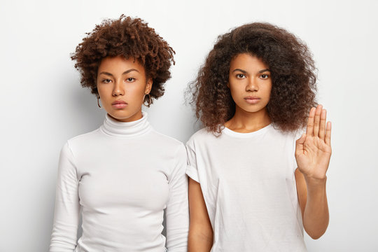 Photo of serious two Afro women have bushy curly hair, one female makes stop gesture, raises palm in rejection, refuses join company, look straightly at camera, stand against white background