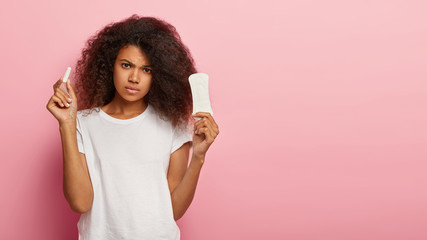 Serious unhappy curly haired lady holds tampon and sanitary pad, thinks which kind of protection better to choose during menstruation or critical days poses on pink wall copy space. Gynecology concept - obrazy, fototapety, plakaty