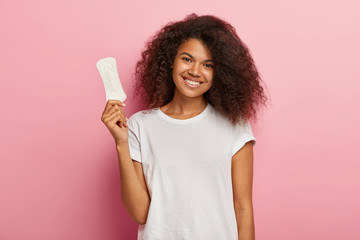 Glad attractive curly female model holds thin sanitary pad, uses feminine intimate product during menstruation or periods, cares about hygiene, wears casual t shirt, models over pink background - obrazy, fototapety, plakaty