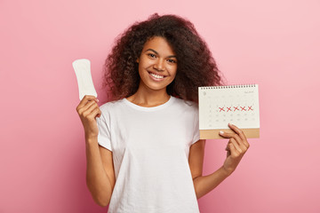 Portrait of good looking feminine girl has Afro hairstyle, holds monthly calendar, clean sanitary napkin, has menstrual cycle, happy to have good women health. People, gynecology, hygiene concept © WHstudio Leushin N