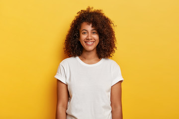 Headshot of beautiful dark skinned curly has pleased expression, rejoices success, enjoys spare time, wears casual t shirt, isolated on yellow background. People, positive emotions, feelings concept - obrazy, fototapety, plakaty