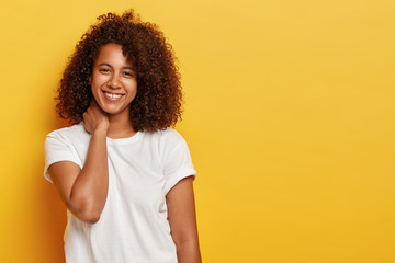 Curly lovely female touches neck, grins joyfully, has flirty look, enjoys spare time, wears casual white t shirt, talks casually with someone, expresses positive emotions isolated on yellow background - obrazy, fototapety, plakaty