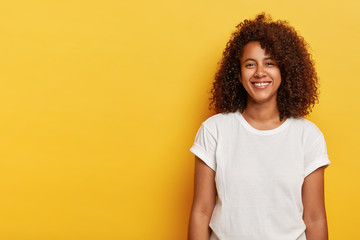 Relaxed female model with curly Afro hairstyle, grins from happiness, glad to have successful day, looks straightly at camera, wears white t shirt, laughs indoor over yellow background, copy space