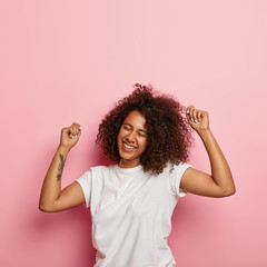 Wellbeing and joy concept. Vertical shot of satisfied carefree curly woman raises tattooed arms,...