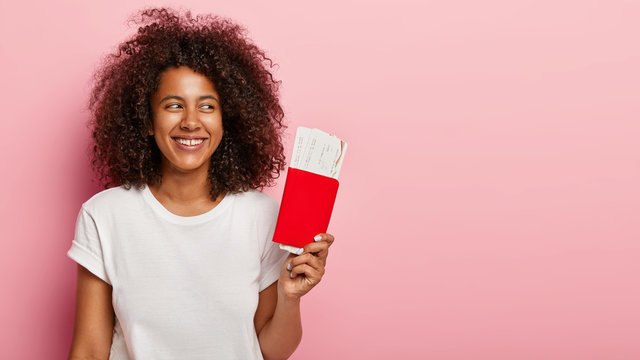 Happy Young Girl With Afro Hairstyle, Holds Passport And Airline Tickets, Rejoices Future Trip On Summer Vacation, Looks Aside Happily, Isolated On Pink Background. Traveller With Documents For Flight