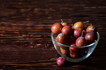 Ripe sweet plum fruits in glass bowl near with scattered plums on dark moody wood table background, hard light, copy space