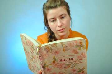 A girl reads the Bible while sitting on a chair on a blue background. The inscription in Russian - "Bible" on the book.