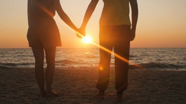 silhouette of a romantic young couple standing on the seashore, man comes up and takes the girl's hand and together enjoying sunrise on the beach, summer vacation and love