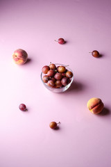 Ripe sweet plum fruits in glass bowl near with scattered plums and peaches on pink background, soft light 