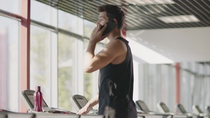 Handsome man using mobile phone in gym. Strong sportsman jogging on treadmill