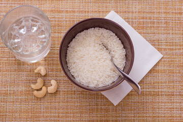 Raw white rice in cup, cup of water, cashew nuts and silver spoon on napkin on gunny background on top with