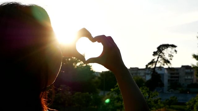 Female hands making heart shape gesture holding sun flare with fingers in the blue sky
