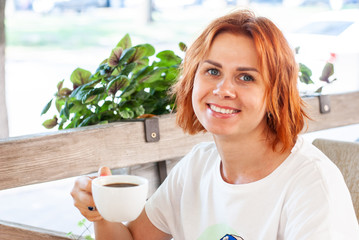 A young girl with red hair sits at a table with a cup of black coffee on a cafe summer platform.
