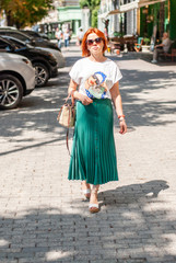 A young girl with red hair in a white T-shirt and a bright green skirt in sunglasses from the sun on her face is walking along the city street.