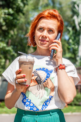 A young girl with red hair in a white T-shirt and a bright green skirt speaks on her cell phone, holds cold coffee in a plastic glass in her hand.