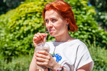 Fototapeta premium A young girl with red hair in a white T-shirt and a bright green skirt drinks cold coffee from a plastic glass through a straw in a city park.