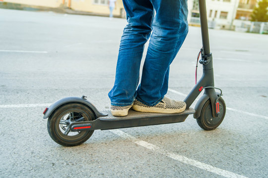 Close Up Of Senior Man Legs In Jeans Riding Electric Kick Scooter On The Yard Asphalt In Summer Day In Front Of The Building