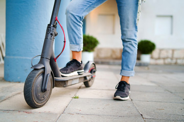 Close up on woman legs feet standing on the electric kick scooter on the pavement wearing jeans and sneakers in summer day