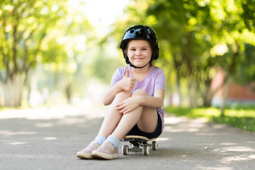 Little girl in a helmet sits on a skateboard and shows a class sign