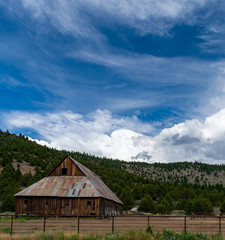 An ancient, weathered barn with rusted metal roof is backed by a forested hill under a vivid blue sky with billowing white clouds.