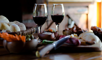 Traditional finger food, which includes different hams, cheeses, vegetables, bread and two glasses of red wine, all arranged on a brown wooden table.