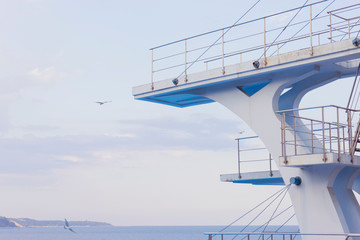 White diving board or tower against a clear blue sky. In the background the sea.