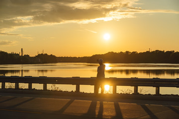Fototapeta premium Silhouette of man sitting on a roadside looking out on a a lake with sunset.