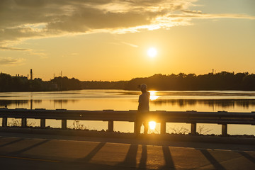 Silhouette of man sitting on a roadside looking out on a a lake with sunset.