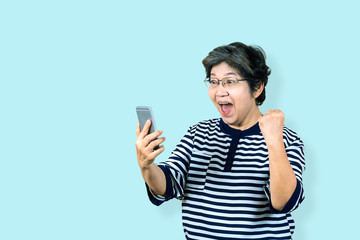 Cheerful senior asian woman holding and looking smartphone on isolated background feeling win ,celebrating and victory. Older female lifestyle concept  blue background.