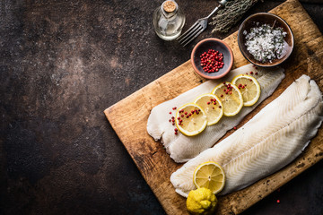 Raw cod fish fillet with lemon slices and herbal salt on rustic wooden cutting board on dark...