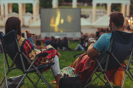 couple sitting in camp-chairs in city park looking movie outdoors at open air cinema