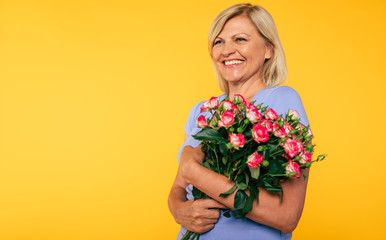 Beautiful lovely smiling blonde senior woman with bouquet of flowers looks on camera and has a good mood while stands over yellow wall. Holidays concepts. Happy mother or grandmother 