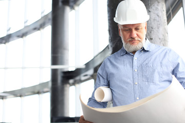 Confident mature architect in corporate suit and hardhat holding a blueprint and looking at it.