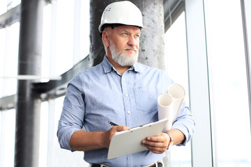 Confident mature business man in hardhat holding blueprint and looking away while standing indoors.