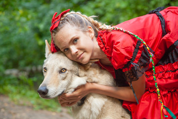 Cheerful pretty young woman in red dress sitting and hugging her wolf dog in the forest