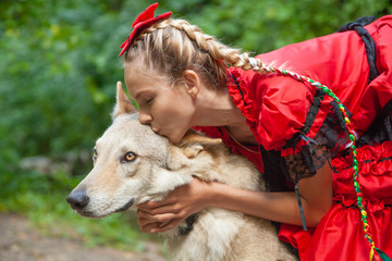 Cheerful pretty young woman in red dress sitting and hugging her wolf dog in the forest