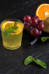 Lemonade drink of soda water, lemon and mint leaves in glass on the black background. Bunch of grapes and sliced citrus fruits
