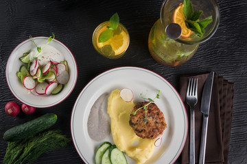 Minced meat cutlets with mashed potatoes on white plate over black wooden background. salad with radish and cucumber. Lemonade drink of soda water