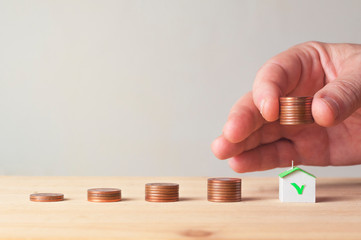 Male hand lift a pile of coin over a paper house with green roof next to other coin piles: saving...