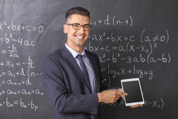 Handsome math teacher with tablet computer near blackboard in classroom