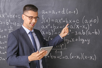 Handsome math teacher with tablet computer near blackboard in classroom