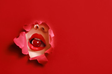 Beautiful young woman with cherry in mouth visible through hole in color paper